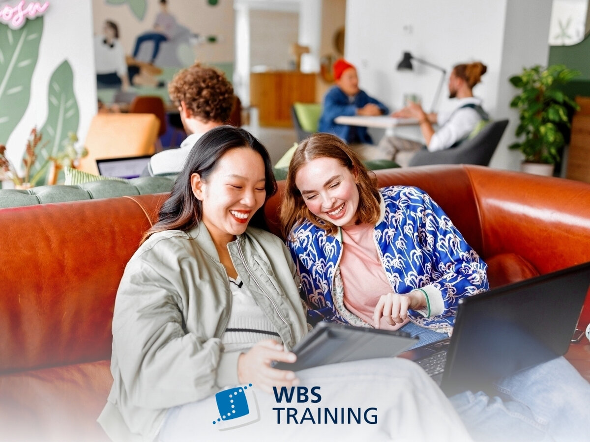Zwei lachende Frauen sitzen auf einer Ledercouch und schauen sich etwas auf einem Tablet an. WBS TRAINING Logo am unteren Bildrand.