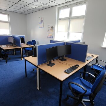 Schulungsraum mit mehreren Arbeitsbereichen, die durch blaue Trennwände voneinander abgegrenzt sind und mit 2 PC-Bildschirmen, Tastatur, Maus und Headset ausgestattet sind. Fensterreihe und Poster an der Wand.