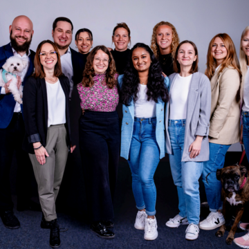 Gruppenbild vom Team. Die Personen lächeln alle freundlich in die Kamera. Ein Mitarbeiter hält einen Hund auf dem Arm.