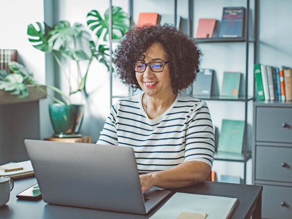 Eine Frau am Laptop in einem Büroraum mit Bücherregal und Pflanze.