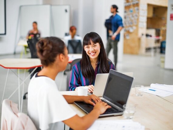 Zwei gut gelaunte Frauen sitzen gemeinsam an einem Tisch vor ihren Laptops.