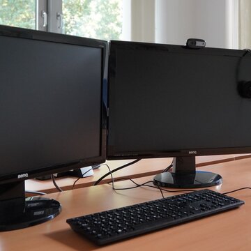 Arbeitsplätze bestehend aus 2 PC-Bildschirmen, Tastatur, Maus und Headset. Im Hintergrund Fenster mit Blick ins Grüne.