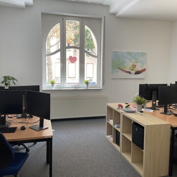Schulungsraum mit mehreren Arbeitsplätzen mit jeweils 2 Bildschirmen, einem Bürostuhl, Tastatur, Maus und Headset. An den Wänden hängen Plakate und Bilder.