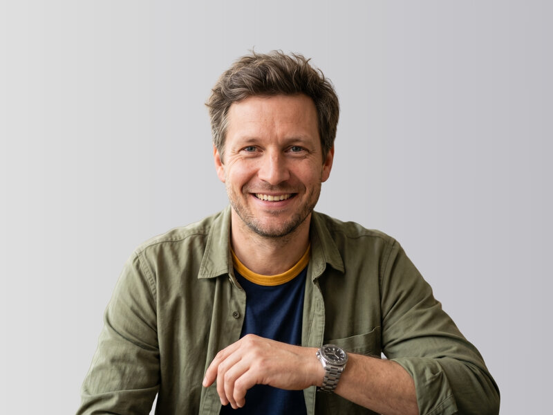Mann mit kurzem braunem Haar und Bart lächelt in grüner Hemdjacke über blauem T-Shirt