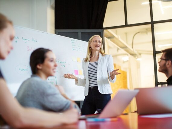 Eine junge, selbstbewusste Frau steht an einem Whiteboard und erklärt ihrem Team ihre Ideen