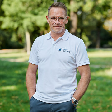 Stefan Schrader steht im weißen T-Shirt mit WBS GRUPPE Logo in der grünen Natur und lächelt in die Kamera.