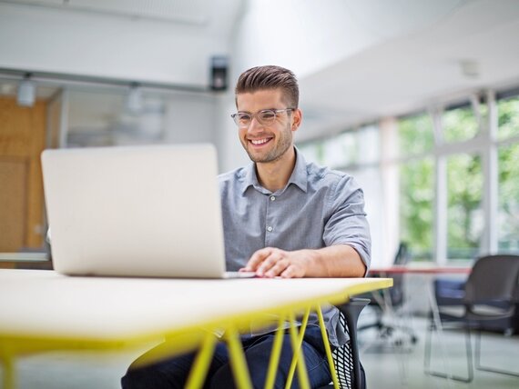 Ein fröhlicher Mann sitz in einem modernen Büro am Schreibtisch und arbeitet am Laptop
