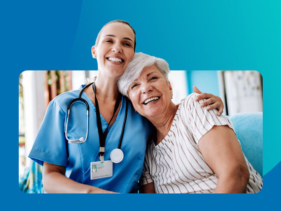 Eine Pflegefachfrau und ihre Patientin sitzen lachend, Arm in Arm auf dem Sofa.