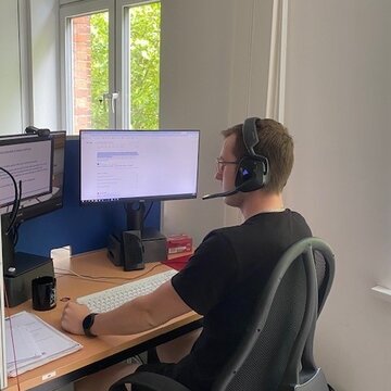 Ein Teilnehmer sitzt an seinem Arbeitsplatz vor 2 PC-Bildschirm und trägt sein Headset.