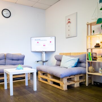 Gemütliche Sitzecke aus 2 modernen Palettensofas mit grauen Kissen, einer Stehlampe, einem Regal und einem Fernseher. Im Hintergrund ein Bild von einem Leuchtturm.