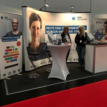 WBS-TRAINING Messestand mit Plakataufstellern und 2 Beraterinnen an einem Stehtisch.