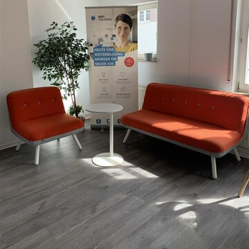 ein Raum mit einem roten Sofa und einem roten Sessel. In der Mitte steht ein kleiner Beistelltisch und neben dem Sessel rechts stehen eine Zimmerpflanze und ein Roll Up Banner der WBS TRAINING
