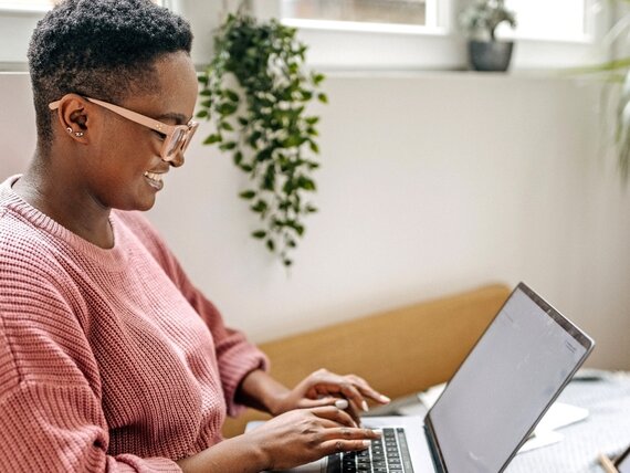 Eine Frau sitzt auf ihrem Bett und arbeitet am Laptop. 