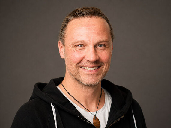 Ein Mann mit Drei-Tage-Bart und schwarzem Hoodie lächelt in die Kamera.