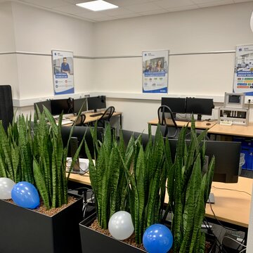 Schulungsraum mit Blick auf die Arbeitsplätze. Im Vordergrund stehen Grünpflanzen mit blau-weißen Ballons.