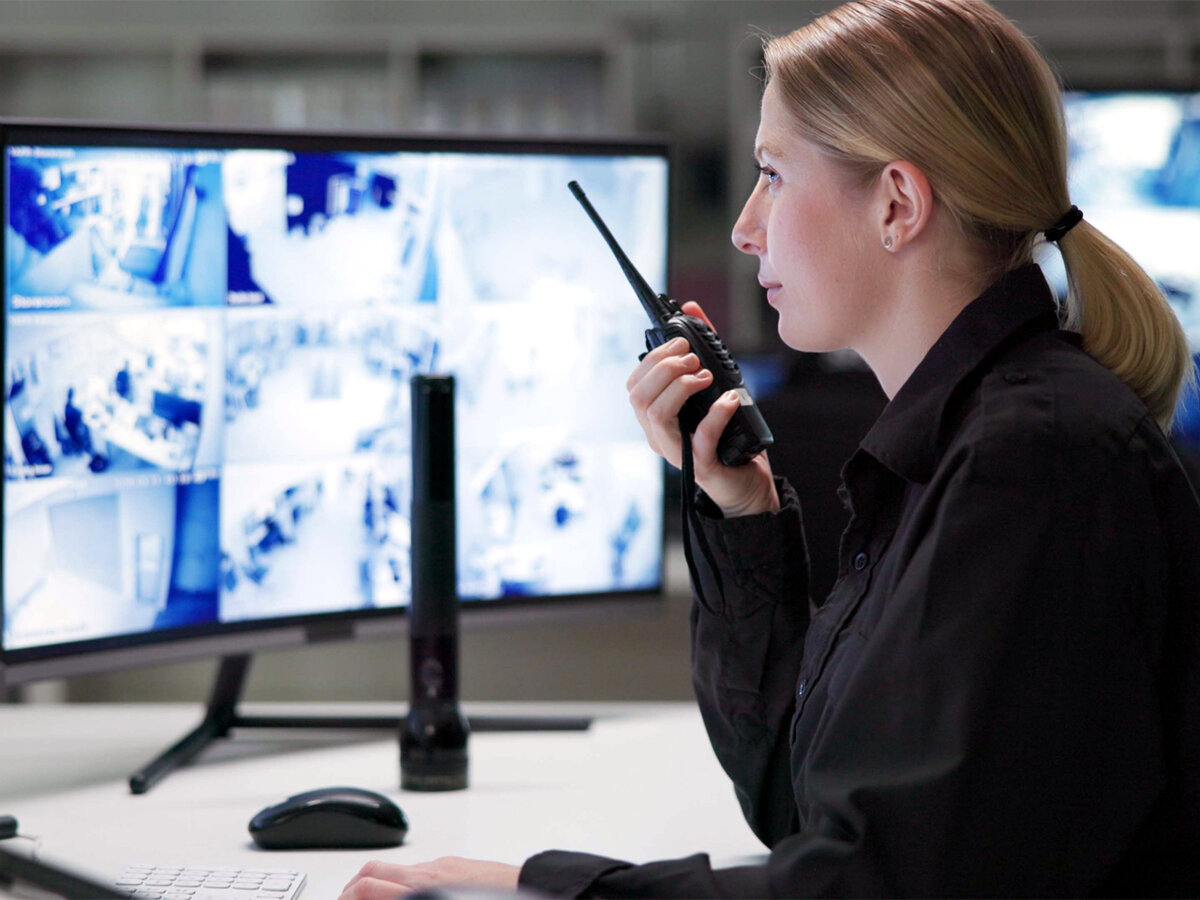 Eine weibliche Wach- und Sicherheitsfachkraft sitzt mit Funkgerät vor zwei Monitoren und überwacht ein Gebäude