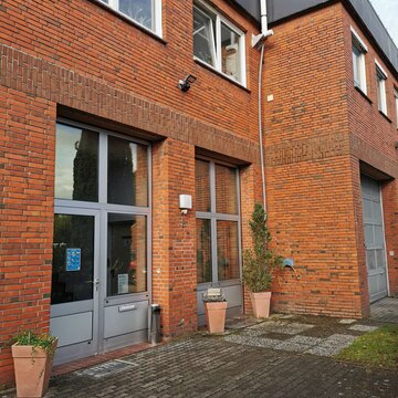 Ein Backsteingebäude mit großen Fenstern und einer Glastür. Vor dem Eingang stehen Blumenkübel mit Pflanzen. Rechts ist ein großes Garagentor zu sehen. Der Boden vor dem Gebäude besteht aus Pflastersteinen und einem kleinen Bereich mit Gras.