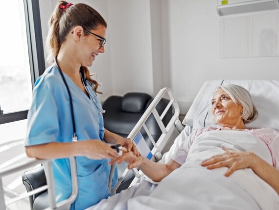 Eine freundliche Pflegerin steht am Krankenbett einer Seniorin und misst ihren Puls