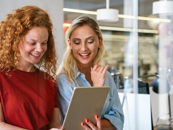 Zwei fröhliche Frauen schauen sich etwas auf einem Tablet an.