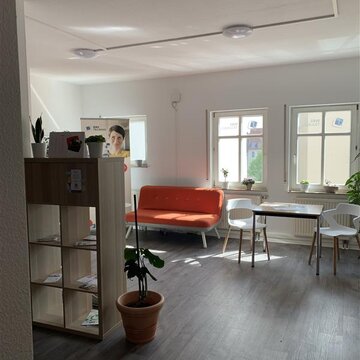 ein Raum mit 3 Fenstern. Links ein Holzregal mit Zimmerpflanzen, dahinter ein rotes Sofa und ein Banner. Rechts neben dem Sofa steht ein Holztisch mit 2 Stühlen.