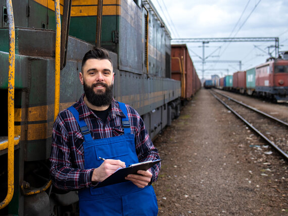 Mann in Arbeitskleidung mit Tablet in der Hand lehnt an einem Rangierzug.