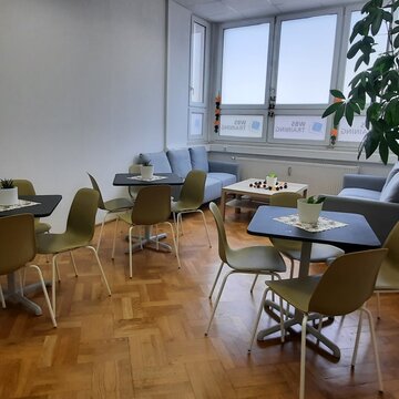 Pausenraum mit Fensterfront und 3 Tischen mit jeweils 4 grünen Stühlen. Im Hintergrund stehen sich 2 Sofas gegenüber, zwischen ihnen ein Beistelltisch. Rechts steht eine große Zimmerpflanze.