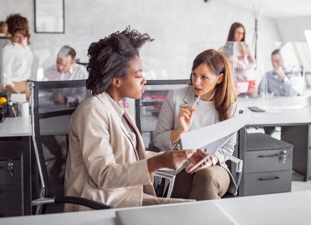 Zwei Frauen sitzen in einem Großraum Büro und besprechen ihre Unterlagen.