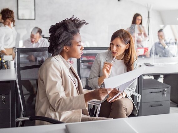 Zwei Frauen sitzen in einem Großraum Büro und besprechen ihre Unterlagen.