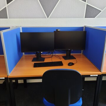 Arbeitsplatz bestehend aus 2 PC-Bildschirmen, Tastatur, Maus und Headset sowie einem blauen Bürostuhl und blauen Trennwänden.