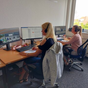 2 Teilnehmerinnen sitzen an ihren Schreibtischen mit zwei Bildschirmen, Maus, Tastatur und Headset