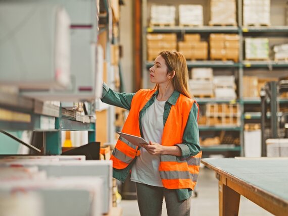 Eine Frau mit Schutzweste und Tablet arbeitet in einem Lager.