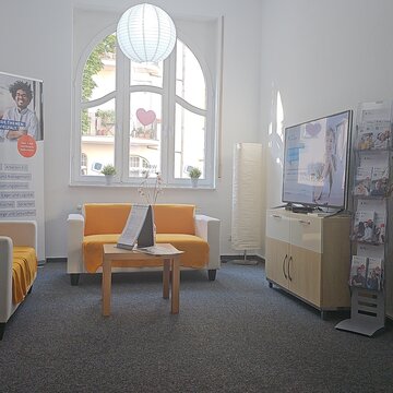 Aufenthaltsraum mit 2 Sofas und einem Beistelltisch. Dahinter ein Roll Up Banner, rechts daneben eine Stehlampe. Rechts ein Sideboard mit TV, daneben ein Flyerständer und eine Pflanze.