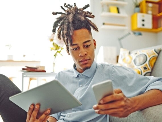 Ein Mann mit Tablet und Smartphone in der Hand sitzt entspannt auf einem Sofa im Homeoffice.
