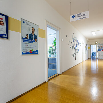 Flur mit mehreren Plakaten an den Wänden und Türen die in verschiedene Zimmer führen. Am Ende des Flures steht ein Roll up Banner und an der Decke hängt ein Schild mit Wegpfeil zum Empfang.