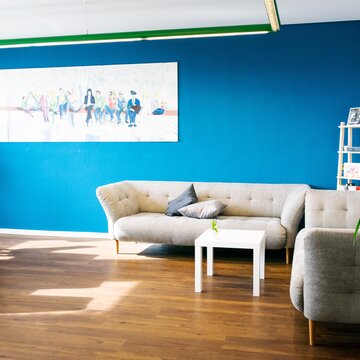 Heller und gemütlicher Aufenthaltsraum mit grauen Sofas, Beistelltisch, Tisch mit Stühlen und einem kunstvollem Bild an der Wand.