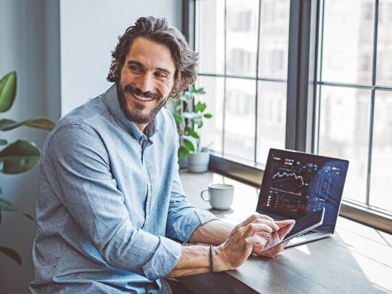 Ein Finanzexperte schaut sich Diagramme und Kurse auf seinem Laptop an und tippt in sein Smartphone