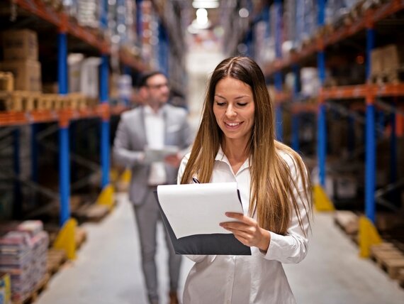 Eine Frau steht in einem Lager und geht ihre Checkliste auf einem Klemmbrett durch.