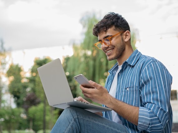 Ein fröhlicher Mann sitzt draußen mit seinem Laptop und Smartphone.