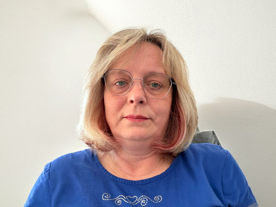Frau mit schulterlangem blonden Haar trägt ein blaues Oberteil und sitzt vor einer weißen Wand