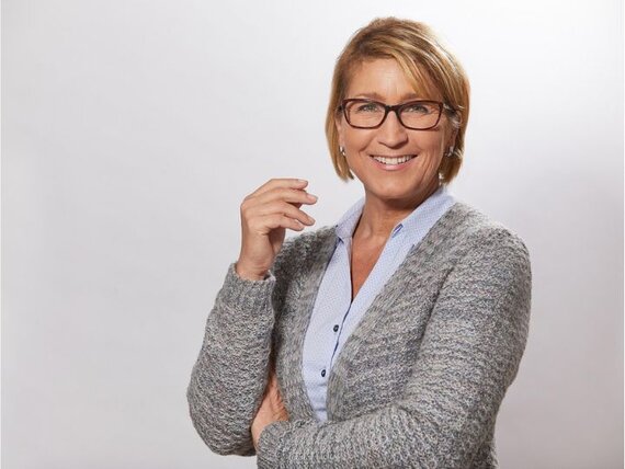 Eine Frau mit Brille und grauer Strickjacke blickt lächelnd in die Kamera.