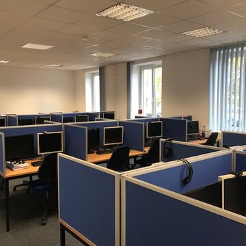 Ein Schulungsraum mit mehreren Arbeitsbereichen, die durch blaue Trennwände voneinander getrennt sind. Jeder Arbeitsplatz besteht aus 2 PC-Bildschirmen, Tastatur und Maus sowie Headset.