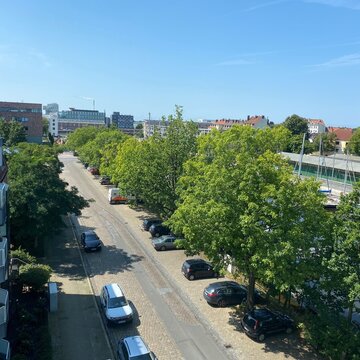 Sicht aus dem Fenster am Standort. Straße mit Fußgängern und Autos und grünen Bäumen. Ein sonniger Tag mit blauem Himmel.