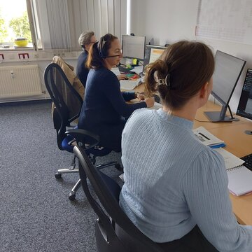 3 Teilnehmerinnen sitzen an ihren Schreibtischen mit zwei Bildschirmen, Maus, Tastatur und Headset