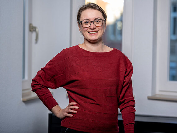 Eine Frau mit Brille in einem roten Pullover. Sie hat den rechten Arm in die Hüfte gestemmt, der linke hängt locker herunter.