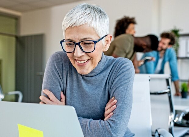 Eine Frau im mittleren Alter sitzt in einem offenem Büro vor ihrem Laptop.