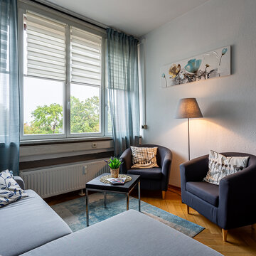Aufenthaltsbereich mit 2 Sesseln und einem hellblauen Sofa. Links befindet sich eine Fensterfront mit blauen Vorhängen und einer Heizung. Rechts steht eine Pflanze. Mittig steht ein Beistelltisch mit Deko und eine Stehlampe.
