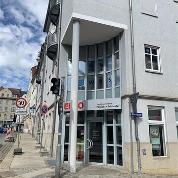 verglaster Haupteingang an der Ecke eines grauen Hauses. Vor dem Eingang steht eine schmale Säule, davor eine Ampel.