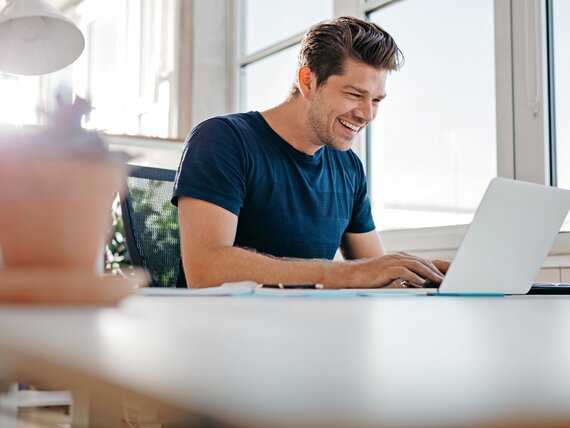 Ein fröhlicher Mann im Homeoffice bei der Arbeit an seinem Laptop.