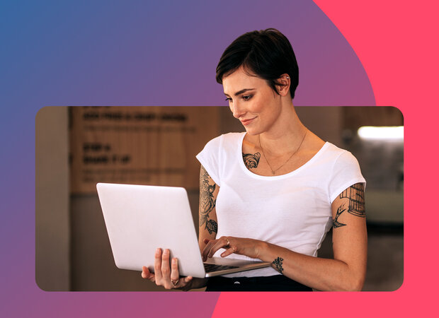 Junge Frau mit weißem T-Shirt und Tätowierungen auf dem Arm schaut auf den aufgeklappten Laptop, den sie auf dem Arm hält