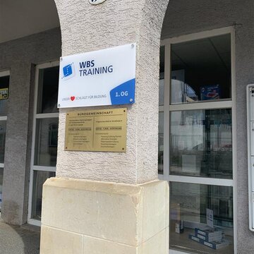 Beschilderung der WBS TRAINING AG an gelber Hausfassade. Im Hintergrund befindet sich ein großes Glasschaufenster.